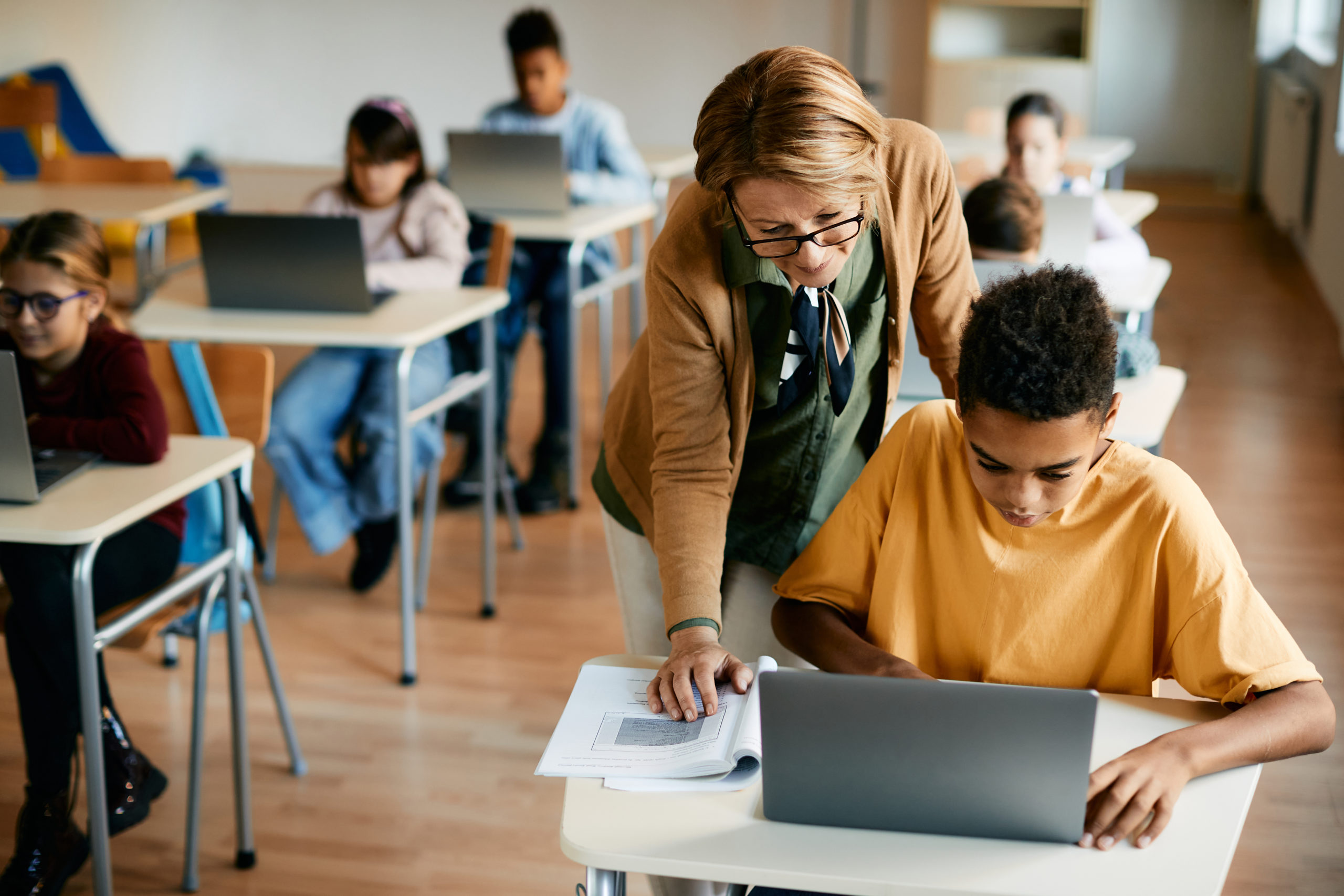 Reife Lehrerin, die ihren Grundschüler bei der Benutzung des Laptops in der Computerklasse in der Schule unterstützt.