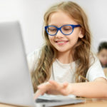Young girl with glasses typing on a laptop with a blurred student in the background.