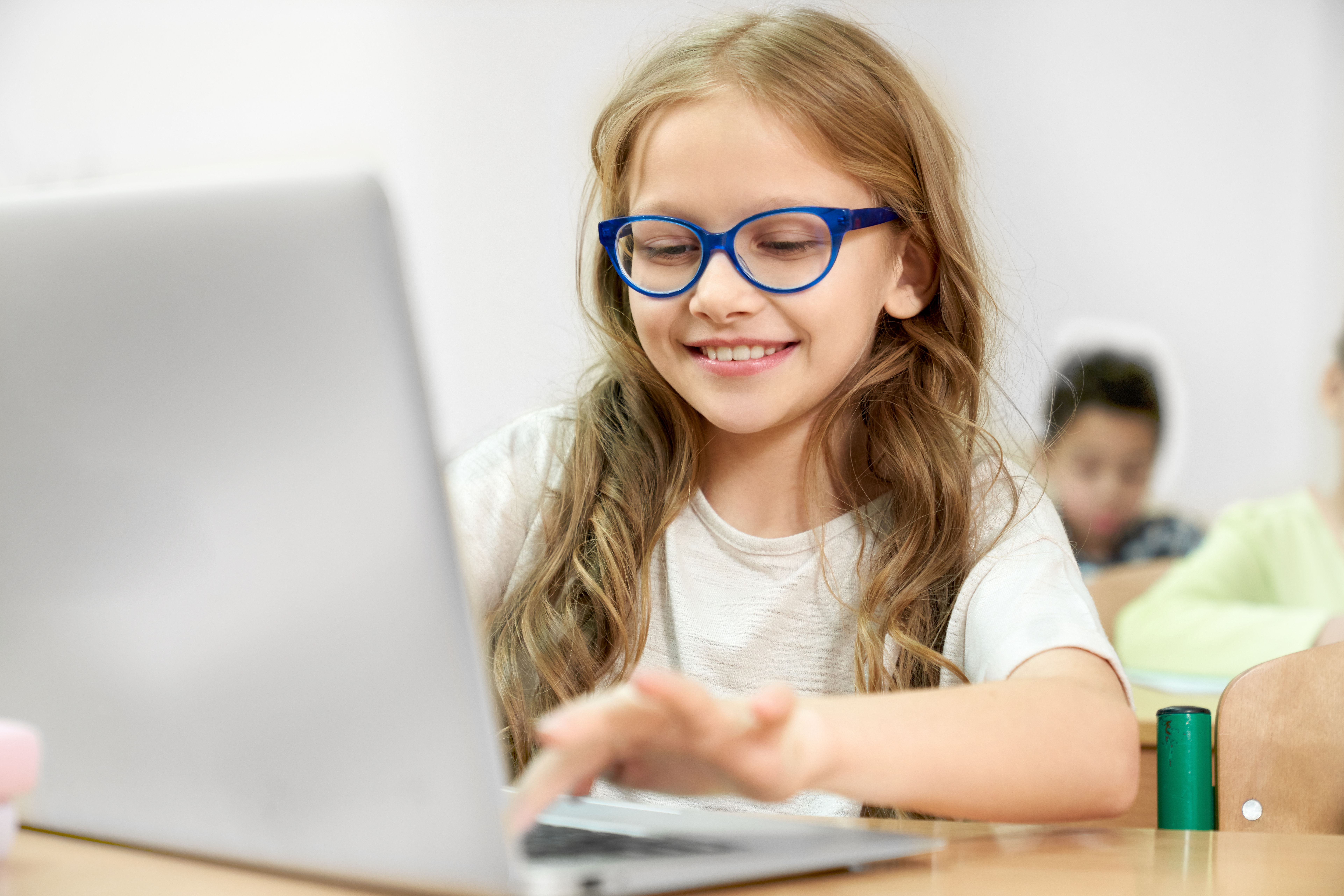 Young girl with glasses typing on a laptop with a blurred student in the background.