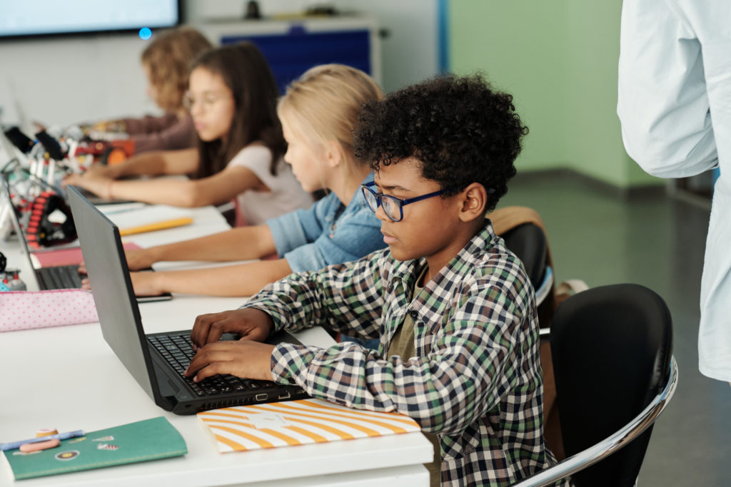 Seitenansicht eines Jungen in einem Klassenzimmer, der an einem Schreibtisch sitzt und einen Laptop benutzt, um eine Lernkontrolle durchzuführen.