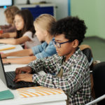 Seitenansicht eines Jungen in einem Klassenzimmer, der an einem Schreibtisch sitzt und einen Laptop benutzt, um eine Lernkontrolle durchzuführen.