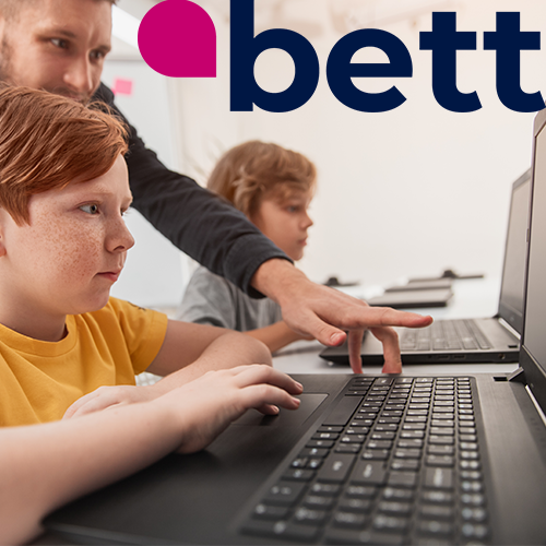 niños sentados ante ordenadores con un hombre mayor detrás señalando la pantalla.