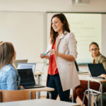 Profesora de pie en un aula sonriendo a los alumnos sentados en sus pupitres con ordenadores portátiles.