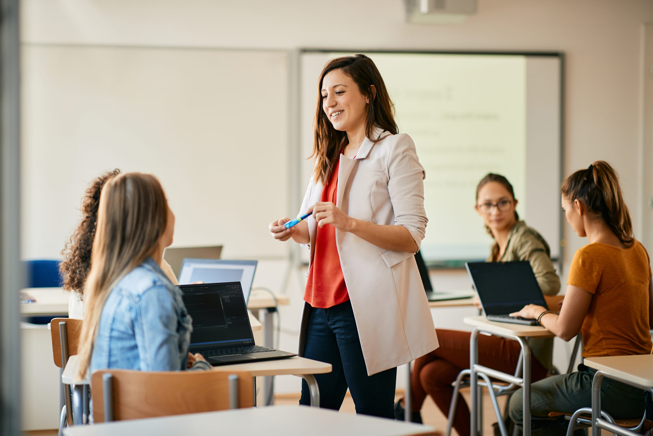 Female teacher standing in a classroom smiling at students sitting at their desks with laptops.