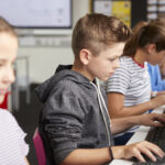 Vista lateral de una fila de alumnos y alumnas sentados ante ordenadores que utilizan herramientas de evaluación estandarizadas.