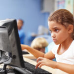 Young schoolgirl in a classroom sitting at a computer and typing on a keyboard participating in an assessment to measure learning loss.