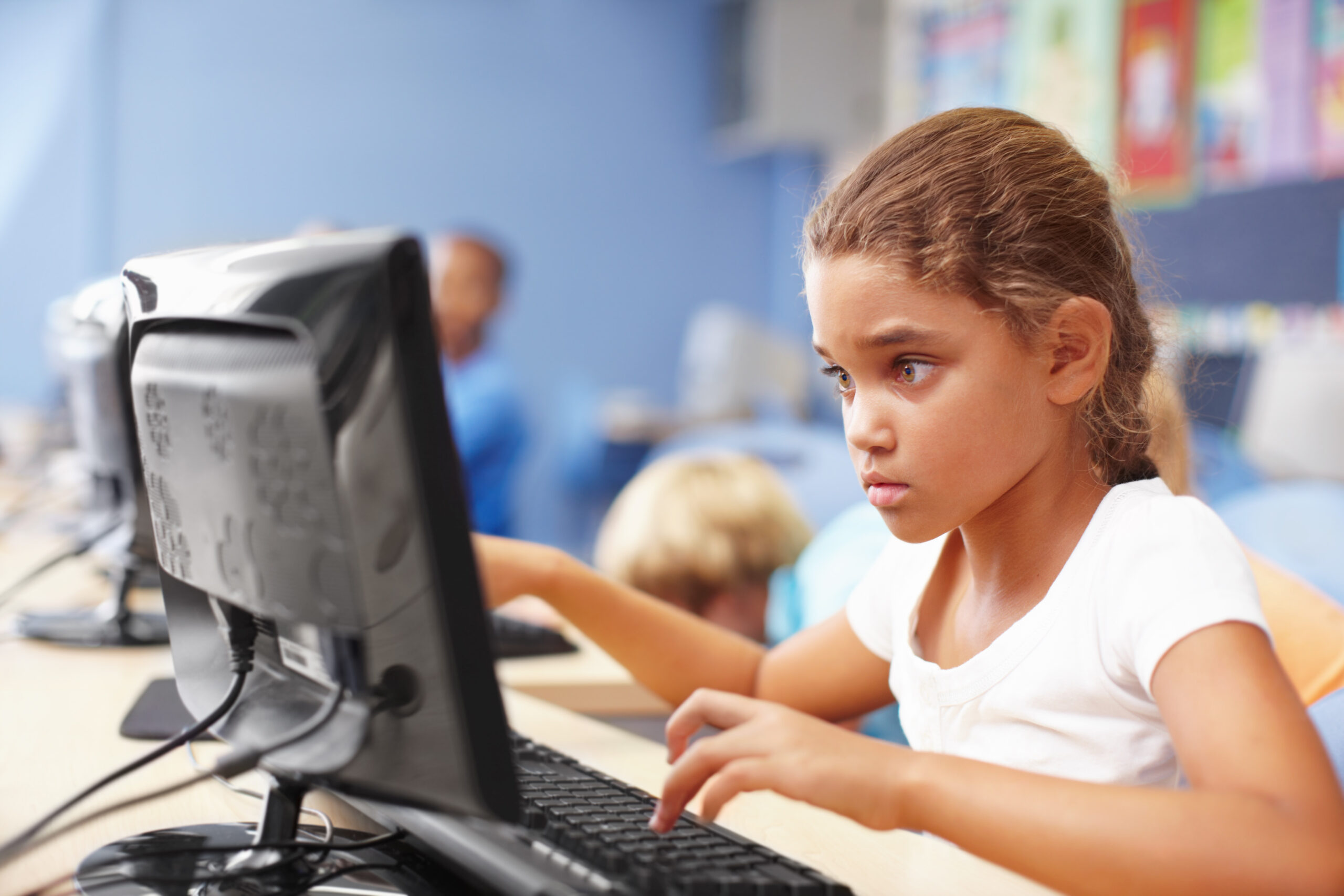 Young schoolgirl in a classroom sitting at a computer and typing on a keyboard participating in an assessment to measure learning loss.