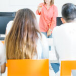 View from behind of students sitting in chairs in a computer lab while looking at a teacher in the front of the class