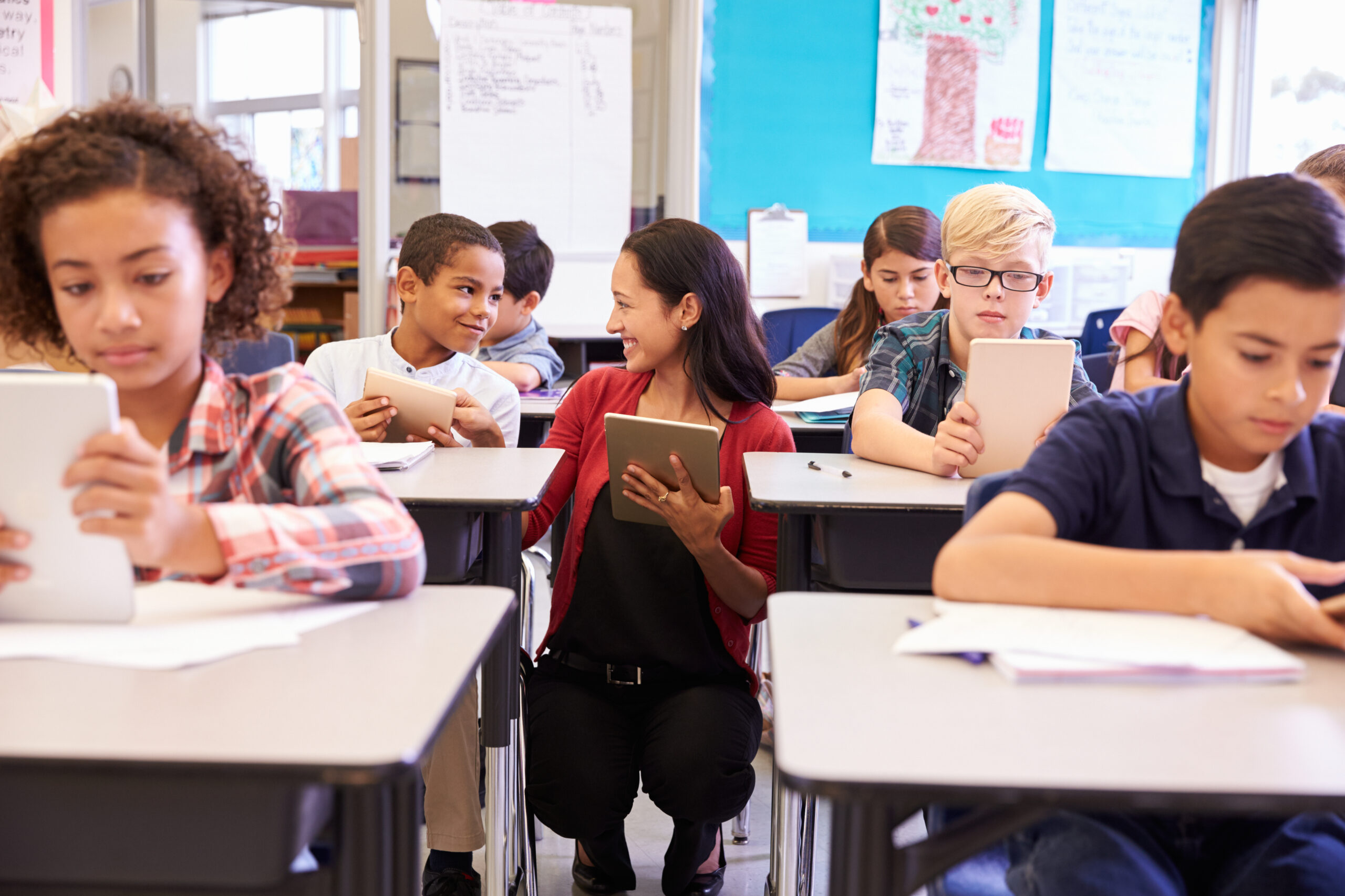 des élèves assis à des bureaux et utilisant des tablettes dans une salle de classe avec un enseignant agenouillé à proximité
