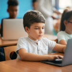 young boy sitting a a desk in a classroom taking a digital exam on a laptop.