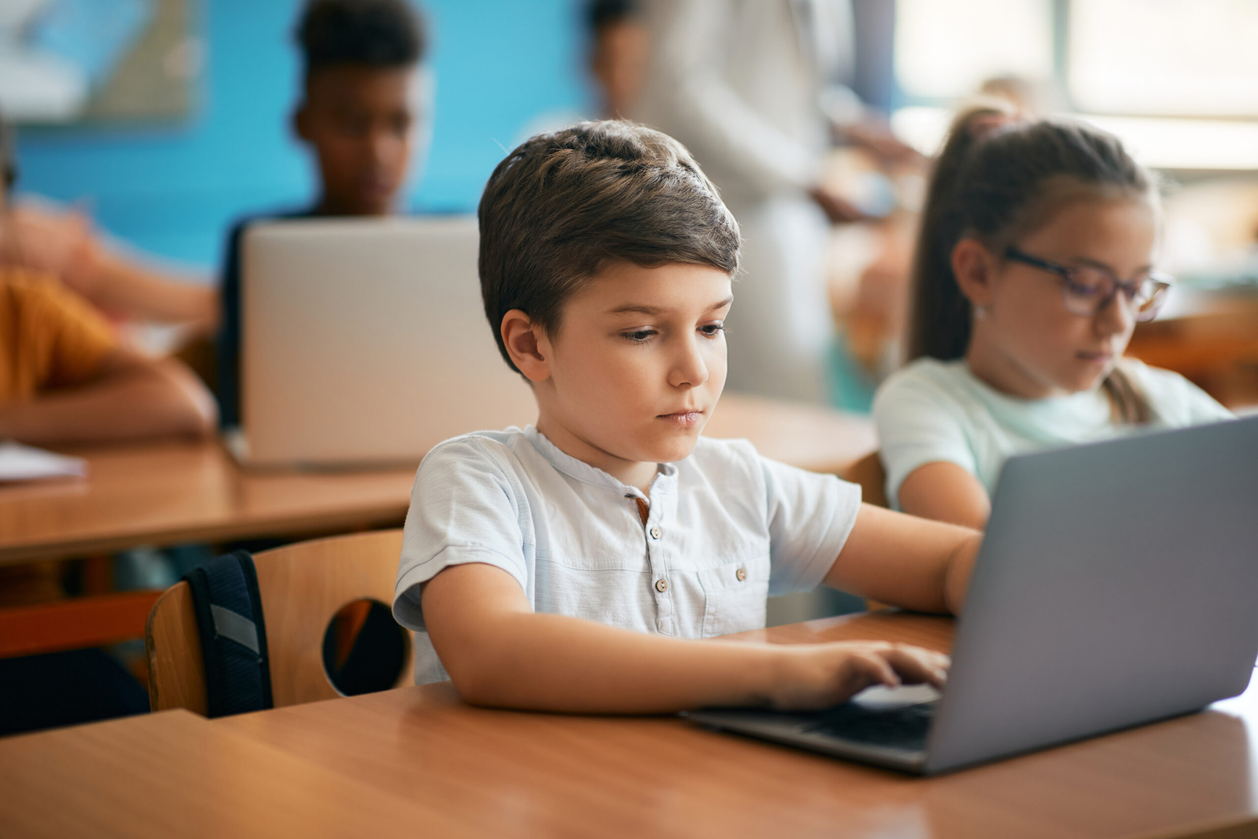 young boy sitting a a desk in a classroom taking a digital exam on a laptop.