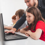 Jeune écolière en cours d'informatique utilisant un ordinateur portable avec un professeur penché sur elle.
