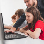 Joven colegiala en clase de informática utilizando un ordenador portátil con el profesor inclinado sobre ella.