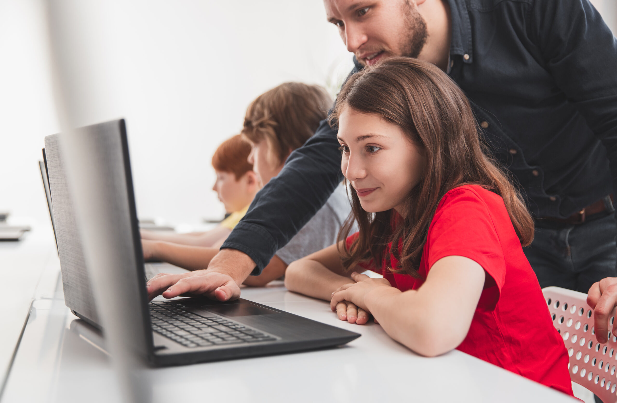 Young schoolgirl in computer class using a laptop with teacher leaning over