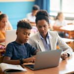 Female teacher kneeling beside a young student using a laptop for digital assessment in the classroom.