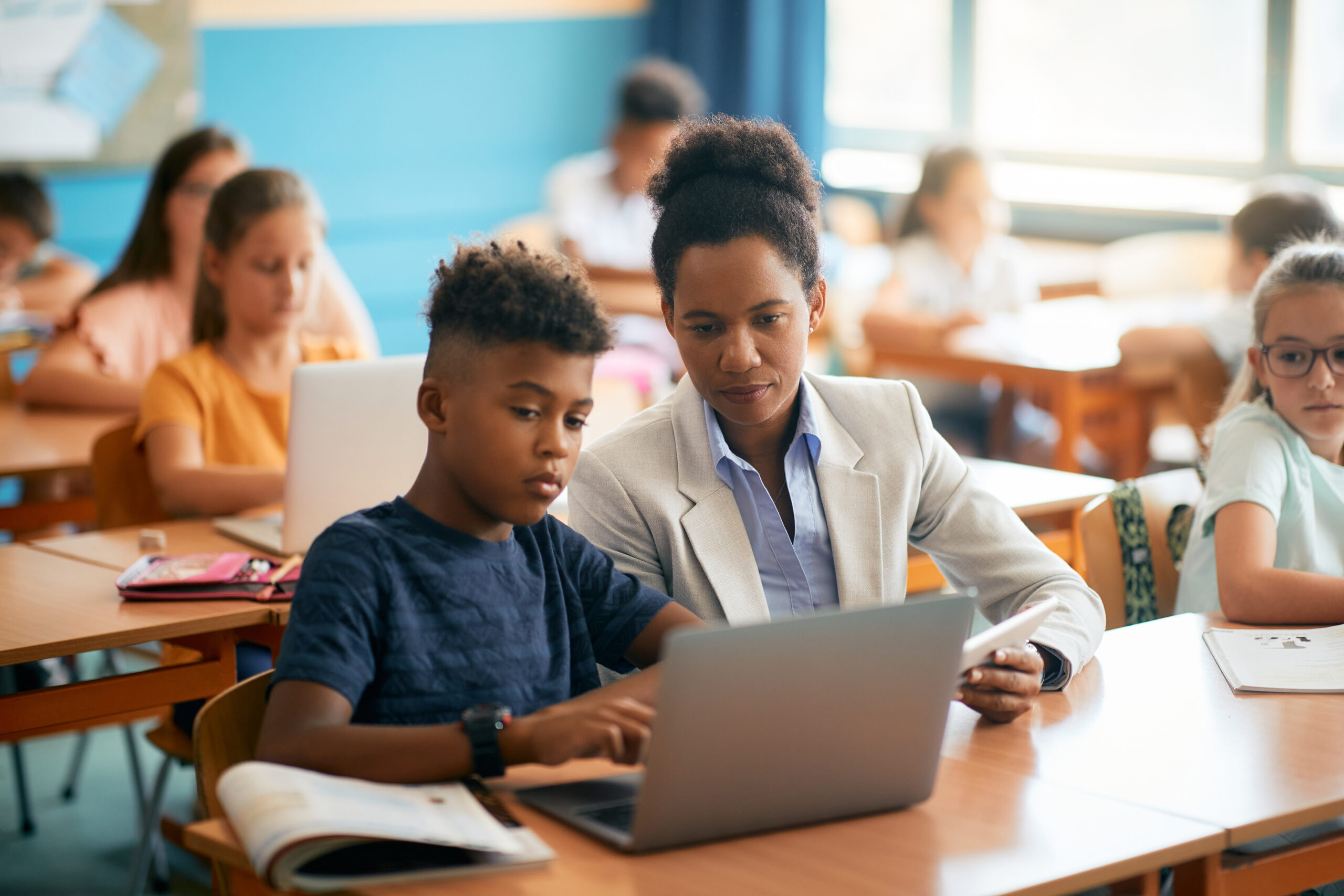 Female teacher kneeling beside a young student using a laptop for digital assessment in the classroom.