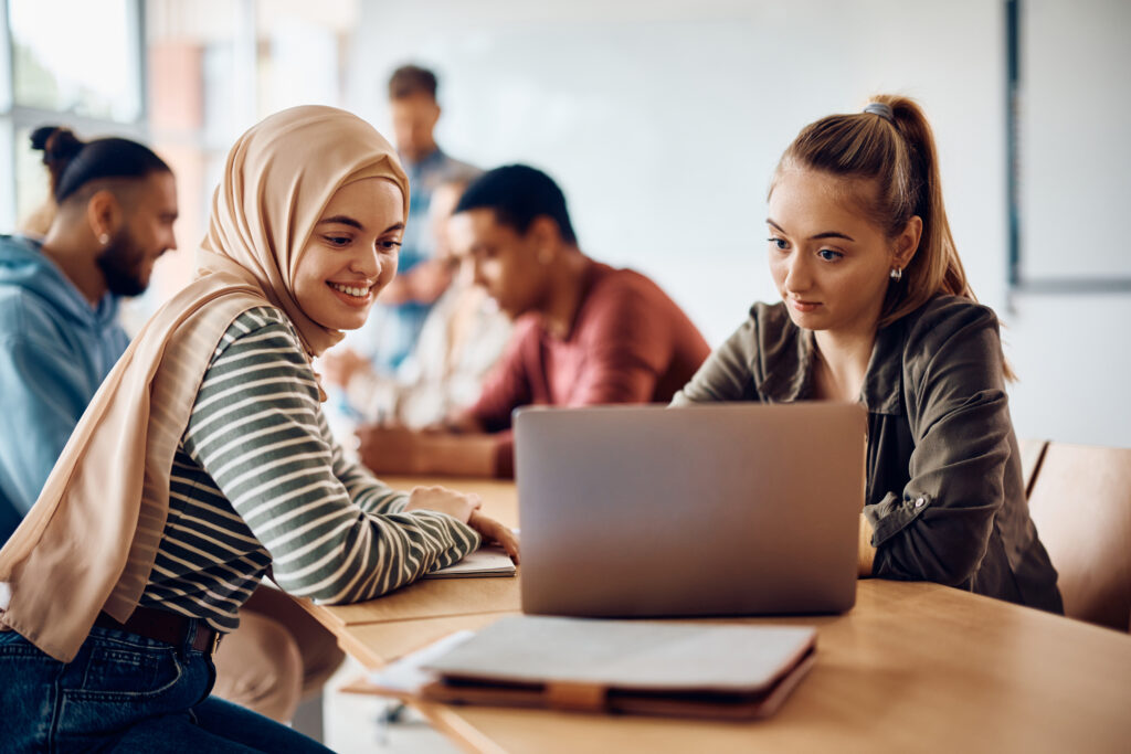 ノートパソコンの画面を見ている2人の女子学生をクローズアップし、背景の他の学生にはピントを合わせず、創造的な評価のアイデアを示す。