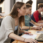 Eine Frau lehnt sich nach vorne und schaut auf einen Computerbildschirm in einem Computerraum, während andere Studenten neben ihr ebenfalls eine sichere Online-Prüfungsplattform nutzen.