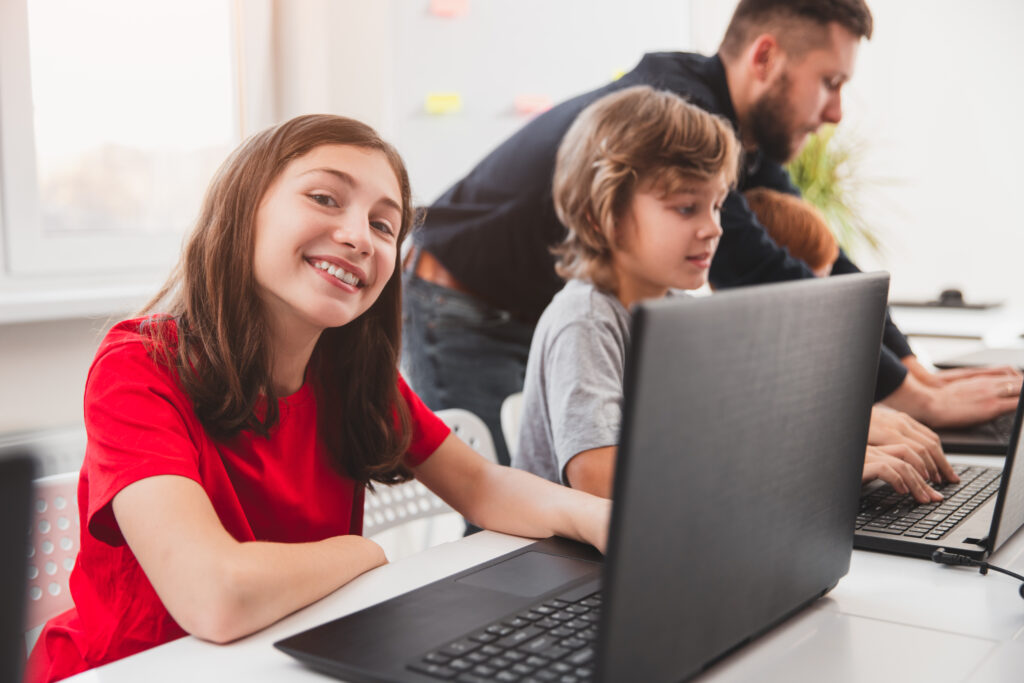 young girl sitting in classroom with her computer next to a young boy and teacher, smiling with her laptop using innovative assessment tools.