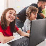 joven sentada en clase con su ordenador junto a un chico y una profesora, sonriendo con su portátil utilizando innovadoras herramientas de evaluación.