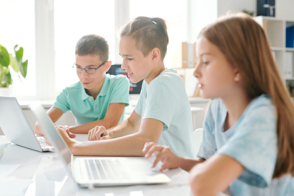 Tres jóvenes escolares con ropa informal sentados en un aula de informática utilizando ordenadores portátiles.