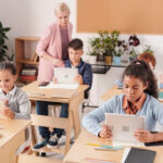 Grupo de escolares contemporáneos con tabletas sentados junto a pupitres en dos filas utilizando herramientas de evaluación educativa y de tecnología educativa como tabletas mientras el profesor consulta a uno de ellos.