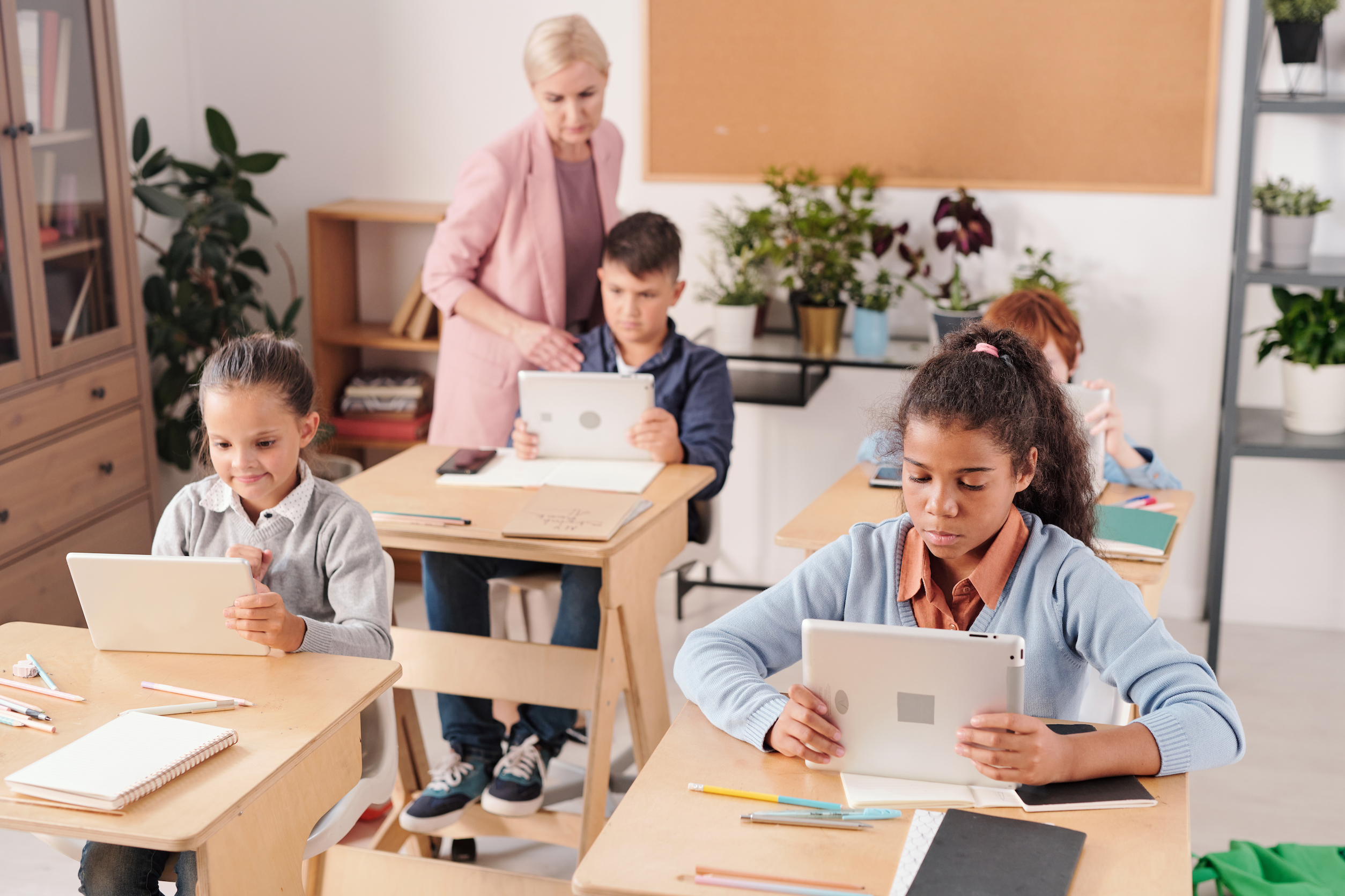 Grupo de escolares contemporáneos con tabletas sentados junto a pupitres en dos filas utilizando herramientas de evaluación educativa y de tecnología educativa como tabletas mientras el profesor consulta a uno de ellos.