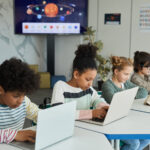 Grupo diverso de niños sentados en fila en el aula de la escuela y utilizando ordenadores portátiles y herramientas de evaluación adaptativa por ordenador.