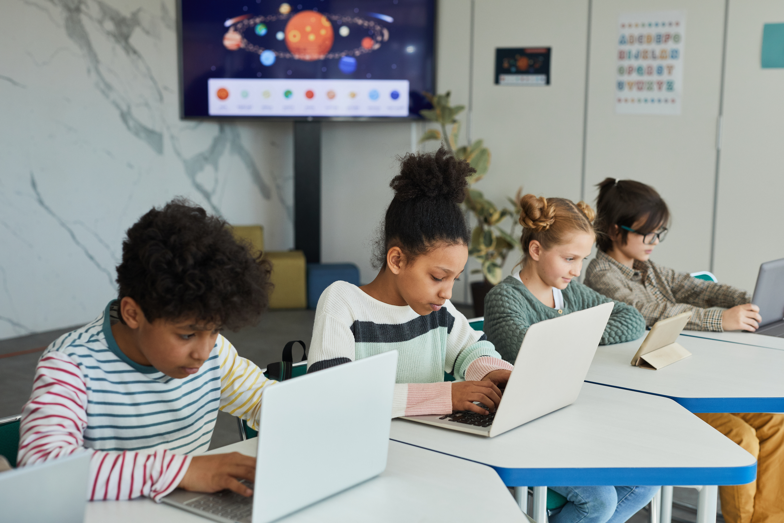 Diverse group of children sitting in row at school classroom and using laptops and computer adaptive assessment tools.