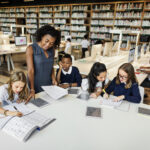 Teaching standing over students sitting at a large desk with open books and tablets on the table, demonstrating literacy assessment and assessment using science of reading strategies