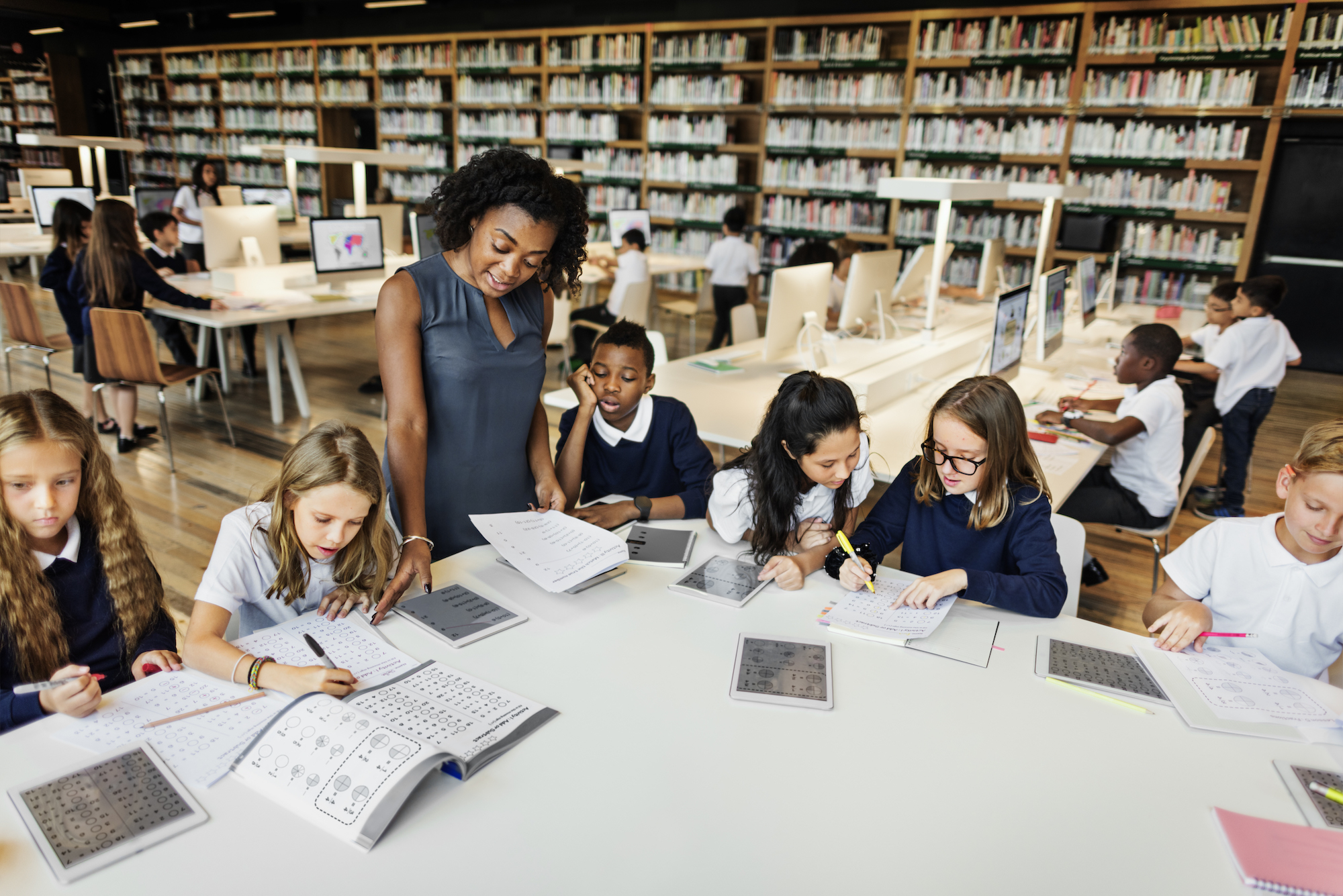 Enseigner debout au-dessus d’élèves assis à un grand bureau avec des livres ouverts et des tablettes sur la table, faire la démonstration de l’évaluation de la littératie et de l’évaluation à l’aide de la science des stratégies de lecture.