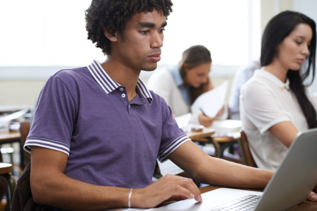 Tシャツを着た若い男性がコンピューターを使用し、焦点が合っていない女性の隣に机に座って、キャリアに即応したスキルを測定するための21世紀の評価の概念を示すクローズアップ
