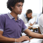 Close up of young man in a t-shirt using a computer and sitting at a desk next to a woman out of focus to demonstrate to concept of 21st-century assessment for measuring career-ready skills