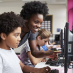 Eine Lehrerin beugt sich über einen jungen männlichen Schüler, der in einer Computerklasse sitzt, die digitale Bewertungsstrategien eingeführt hat.