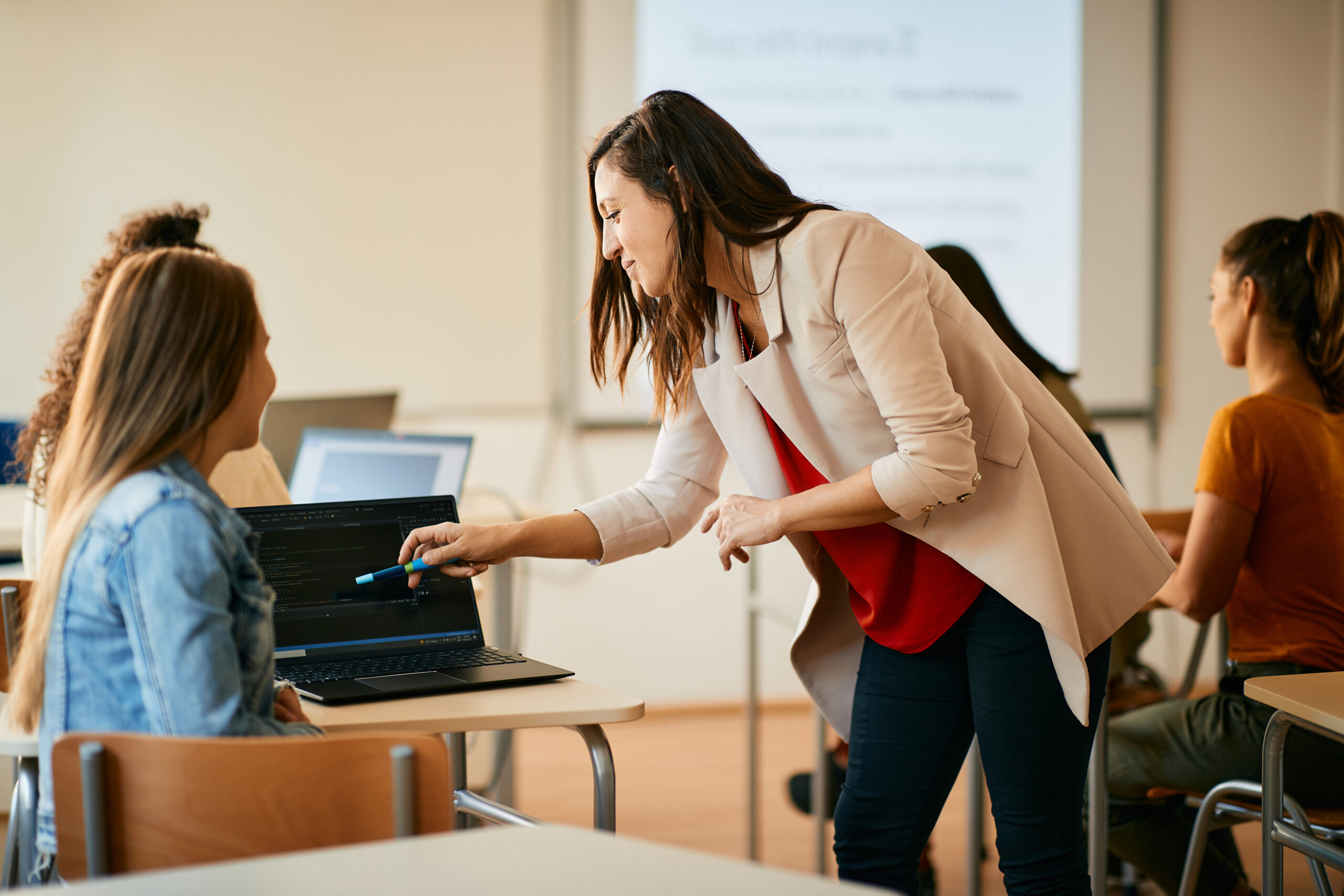 Junge Lehrerin in einem Klassenzimmer, die sich über den Schreibtisch einer Schülerin beugt und auf ihren Computer zeigt, um das Konzept der Benchmark-Bewertungen zu erläutern