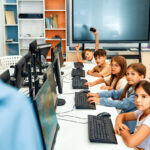 Des écoliers assis en rang dans une classe d'informatique face à leur ordinateur et regardant l'enseignant sur le côté, illustrant le concept d'évaluation globale.