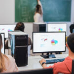 Vue arrière d'élèves en classe, assis à des pupitres devant des ordinateurs, face à une enseignante qui, le dos tourné, dessine sur un tableau, illustrant des élèves en train de passer des évaluations formatives ou sommatives.