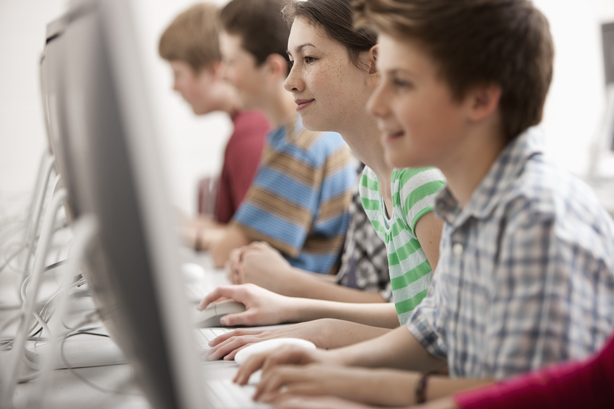 Profile view of four children sitting in a row of desks typing on school computers while taking adaptive tests.