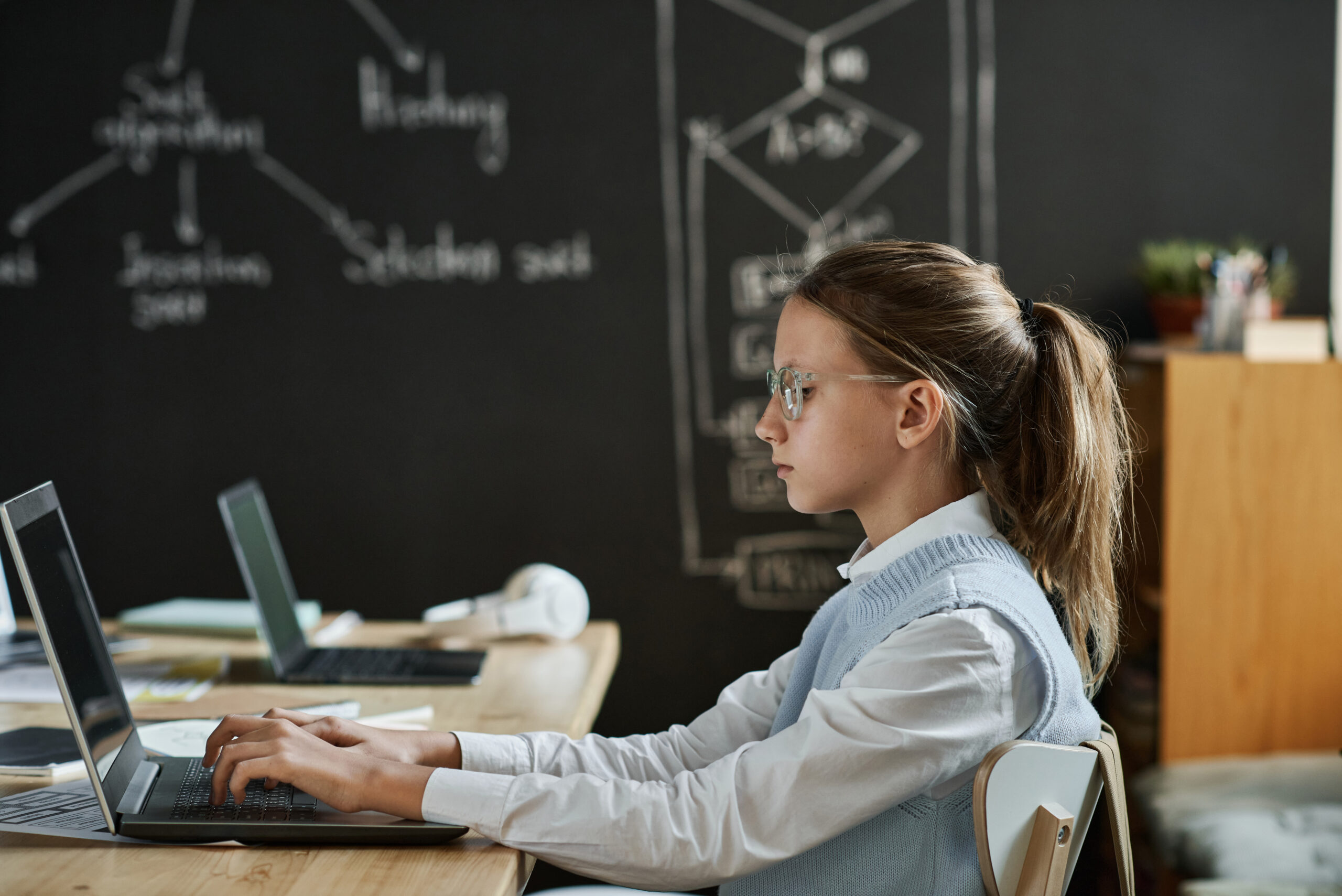 Side view of school girl sitting at a desk in front of a chalkboard facing her laptop.