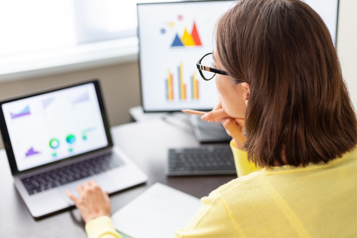 Woman wearing glasses sitting at a desk looking at a laptop and desktop monitor that each show computer-based reports on the screen.