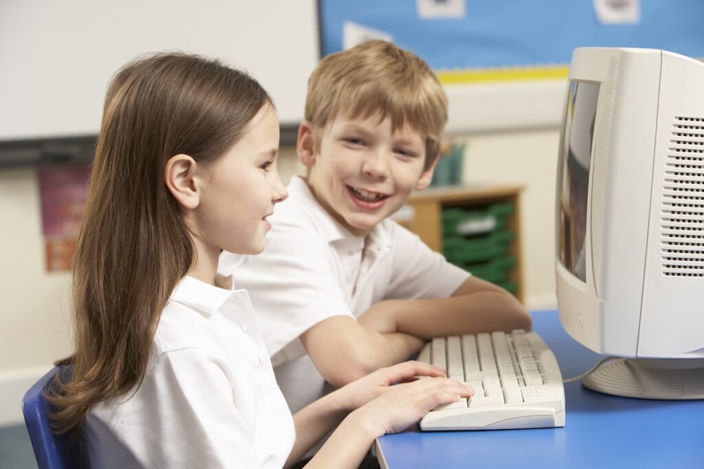 boy and girl schoolchildren sitting and leaning over a desktop computer while smiling.