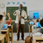 Der Lehrer steht vor dem Klassenzimmer neben einem Fernseher. Die Schüler sitzen dem Lehrer gegenüber und schauen auf die Computer, die das Konzept der Technologie, die offene Standards nutzt, zeigen