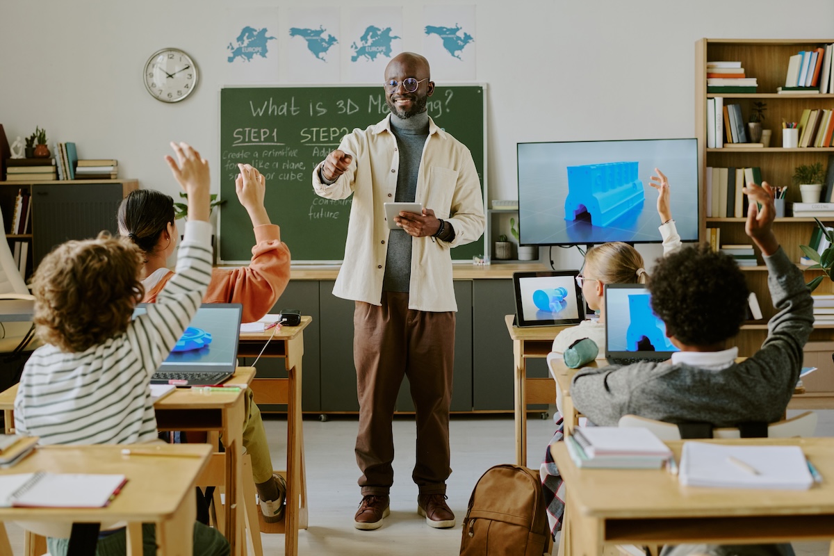 Teacher standing in front of the classroom next to a tv. Students seated facing the teacher looking at computers, showing the concept of technology leveraging open standards