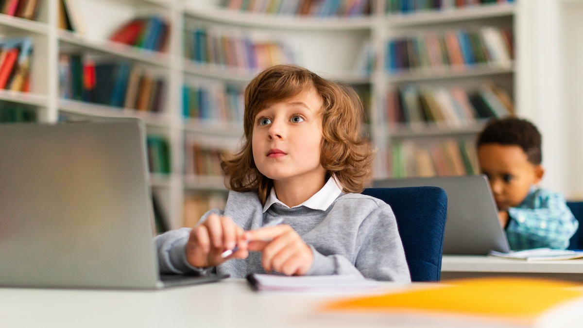 Young boy sitting at a desk in class in front of a computer with a notebook on the desk showcasing an example of student taking a diagnostic reading assessment