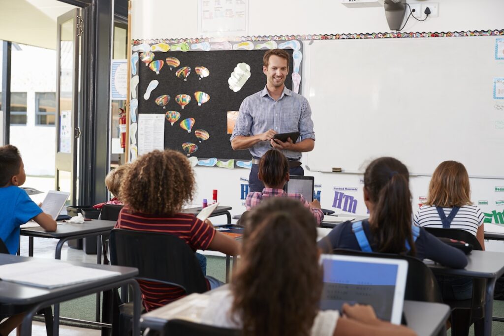 Male teacher standing in front of a classroom reviewing students progress.