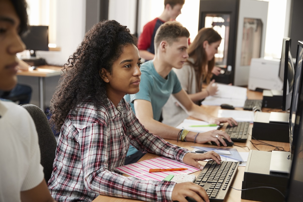 Students sitting in a horizontal row at a classroom table while working at computers, demonstrating the concept of CAT testing