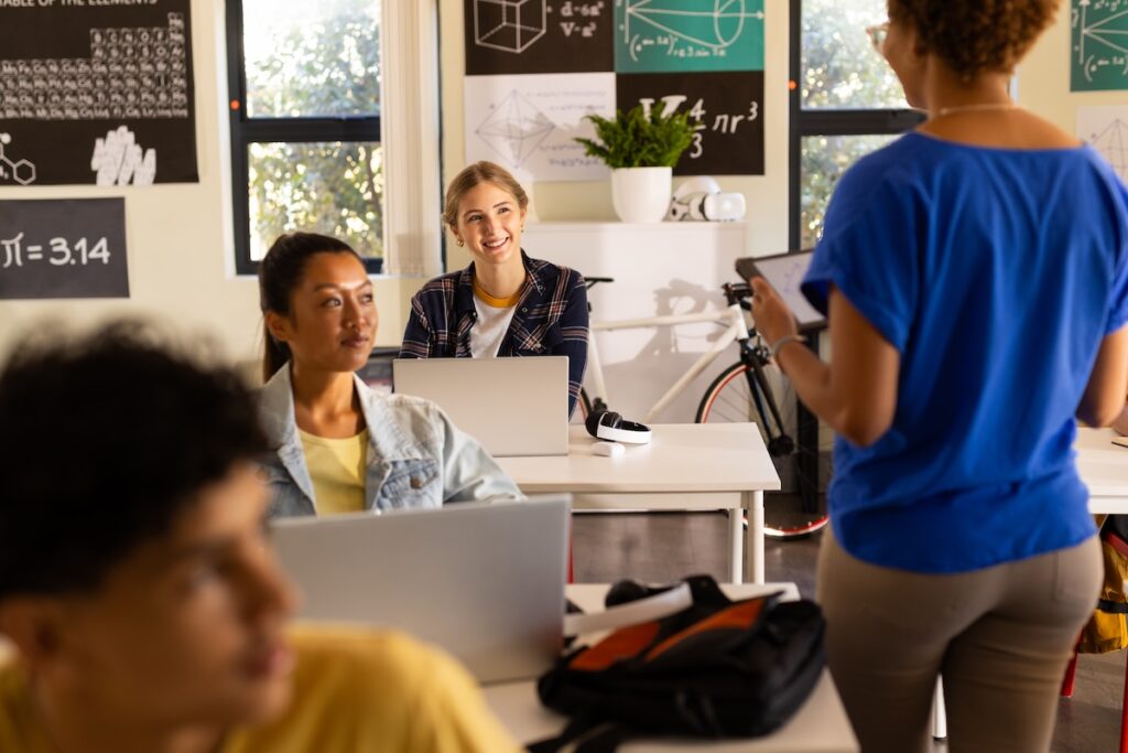 Highschool-Schülerinnen und -Schüler sitzen an ihren Tischen vor ihren Laptops in einem Klassenzimmer, während eine Lehrerin in der Mitte des Raums steht und ein Tablet in der Hand hält, um zu demonstrieren, wie Lehrerinnen und Lehrer die Arbeiten der Schülerinnen und Schüler auf KI überprüfen