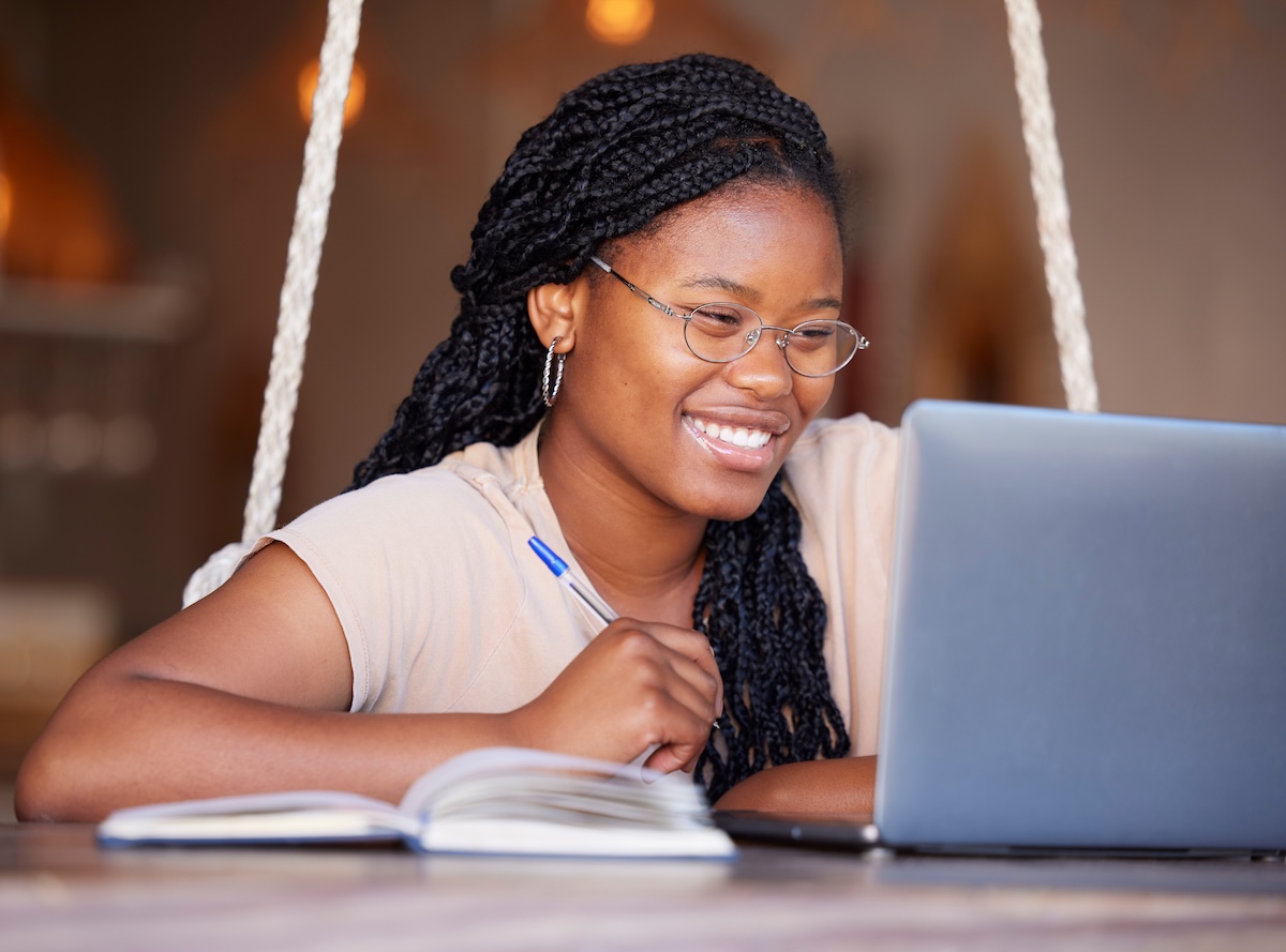 Smiling female college student sitting in front of a laptop while holding a pen and taking notes in a notebook