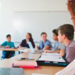 Male high school teacher sitting on a desk teaching as a female student sitting in a semi circle row of desks raises her hand, showing the concept of classroom management strategies for high school
