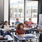 Teacher standing and facing a classroom of elementary students seated at their desks.